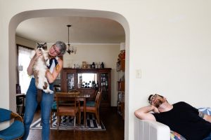 Woman holds up her ragdoll cat at her home while her partner looks on from the couch
